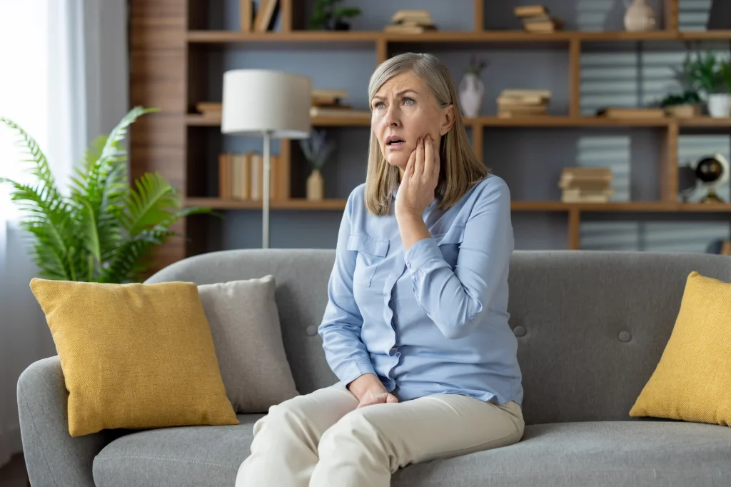 Struggling mature lady feeling dental pain and rubbing cheek with palm while sitting with open